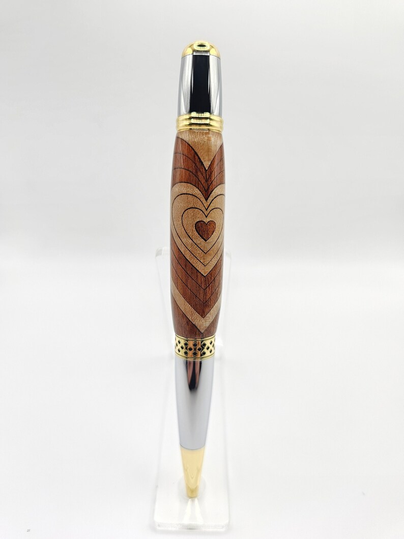 May include: A wooden pen with a heart design in shades of brown and gold. The pen has a silver metal tip and a gold band.