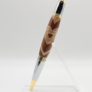 May include: A wooden pen with a silver and gold finish. The pen has a heart design carved into the wood. The pen is displayed on a clear acrylic stand.