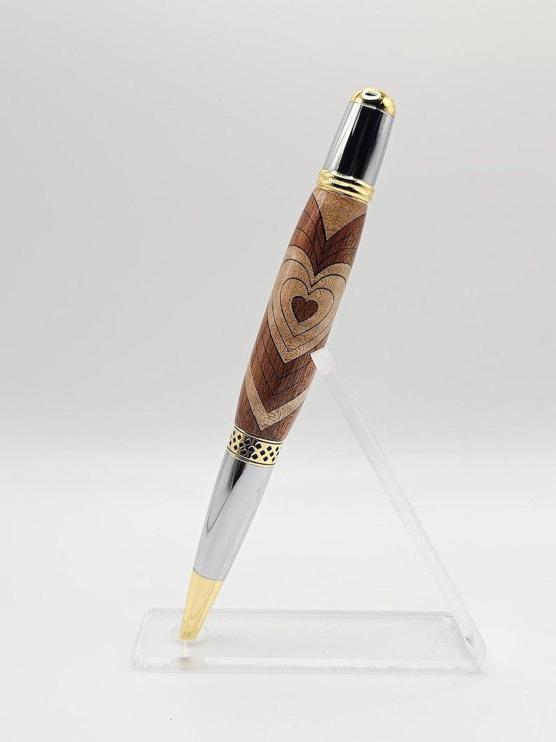 May include: A wooden pen with a silver and gold finish. The pen has a heart design carved into the wood. The pen is displayed on a clear acrylic stand.