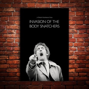 Op de afbeelding: Een zwart-wit poster voor de film "Invasion of the Body Snatchers". De poster toont een man met een geschokte uitdrukking die naar de kijker wijst. De tekst "A Philip Kaufman Film" en "Invasion of the Body Snatchers" is in wit op een zwarte achtergrond gedrukt.