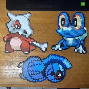 May include: Three pixel art Pokemon characters made with perler beads. The characters are a blue and white frog, a brown and white dog, and a blue and purple creature.