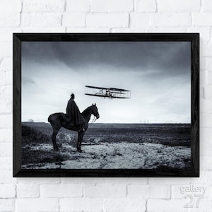 May include: Black and white framed print featuring a person on horseback observing an early biplane in flight. The image captures a historical moment with a vintage aesthetic, set against a field and cloudy sky.