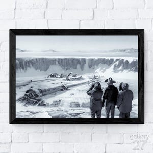 May include: Black and white framed print depicting a snowy landscape with a large, skeletal structure in the foreground. Three figures in winter attire stand facing the scene. The print is framed in a black wooden frame.
