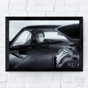 May include: Black and white framed print featuring a man in a classic car. The image shows the driver looking forward through the windshield. The car's engine is visible on the right side of the frame. The print is in a black frame.