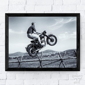 May include: Monochrome framed print showing a man on a motorcycle leaping over a barbed wire fence. The motorcycle's front wheel is off the ground. The background depicts a rural landscape under a cloudy sky.