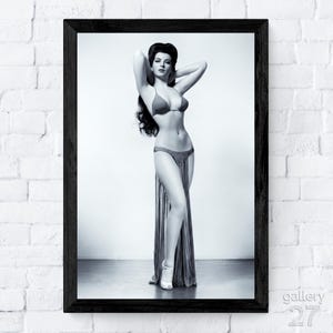 Puede incluir: Impresión enmarcada en blanco y negro de una mujer en bikini y falda larga, posando con los brazos por encima de la cabeza. La imagen es una fotografía de estilo vintage, con las palabras "gallery 27" en la esquina inferior derecha.