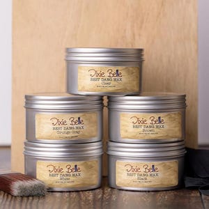May include: A stack of six silver metal tins of Dixie Belle Best Dang Wax in various colors, including Clear, Grunge Gray, Brown, White, and Black. Each tin has a label with the product name and color. A paintbrush is visible on the left.