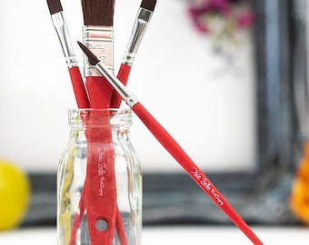 May include: A clear glass jar filled with red-handled paint brushes. The brushes have brown bristles and are of various sizes and shapes. The jar is set against a blurred background with yellow apples and a decorative frame.