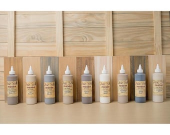May include: A collection of Dixie Belle paint bottles in various neutral colors, including gray, beige, and white, with white applicator tips. The bottles are arranged in a row against a wooden backdrop.