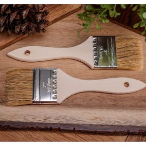 May include: Two paint brushes with light-colored wooden handles and natural bristles. The metal band on each brush is stamped with "2" and "50.8mm". The brushes are displayed on a wooden surface, suggesting a painting or craft theme.