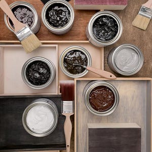 May include: An overhead shot of paint cans and brushes arranged in wooden trays. The cans contain various colors of paint, including black, white, and brown. Paintbrushes with wooden handles are placed near the cans, suggesting a painting or staining project.