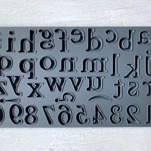 May include: Gray silicone alphabet and number mold with letters a through z and numbers 0 through 9. The mold is on a white wooden surface.