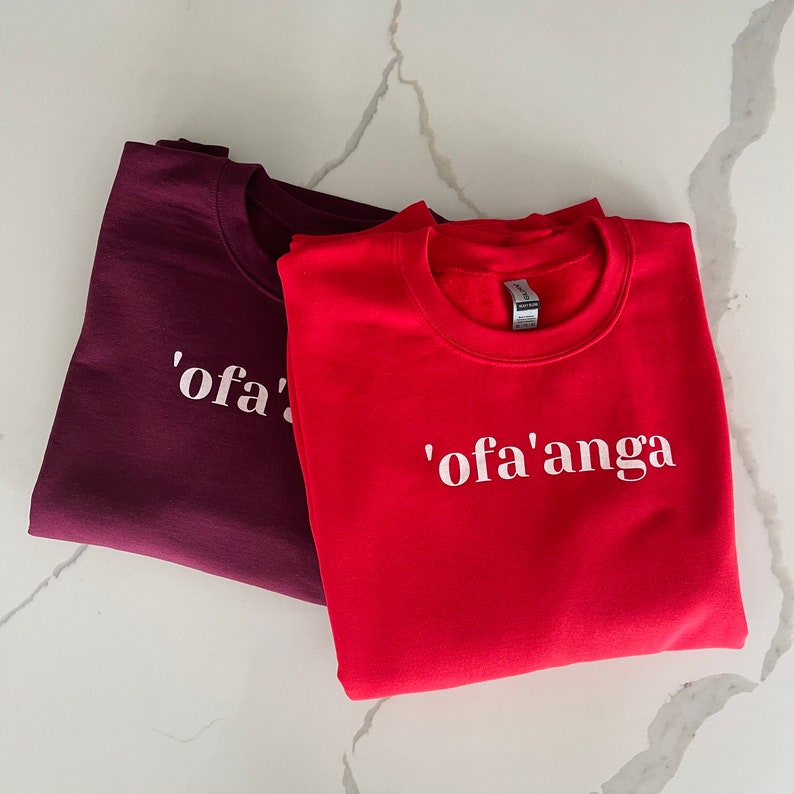 May include: Two crew neck sweatshirts, one burgundy and one red, with white text. The burgundy sweatshirt says "'ofa'" and the red sweatshirt says "'ofa'anga".