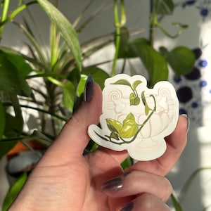 Puede incluir: Una pegatina que representa una urna color crema con una planta verde que crece. La pegatina se sostiene en una mano, con una planta verde en el fondo.