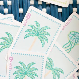 Pode incluir: Um close-up de um baralho de cartas com fundo azul e branco. As cartas apresentam v&aacute;rias ilustra&ccedil;&otilde;es, incluindo palmeiras, um drag&atilde;o e uma flor. As cartas s&atilde;o numeradas com o texto "BAM" e um n&uacute;mero.