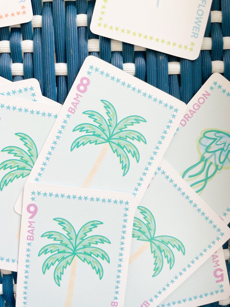 Pode incluir: Um close-up de um baralho de cartas com fundo azul e branco. As cartas apresentam v&aacute;rias ilustra&ccedil;&otilde;es, incluindo palmeiras, um drag&atilde;o e uma flor. As cartas s&atilde;o numeradas com o texto "BAM" e um n&uacute;mero.