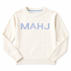 May include: Cream-colored sweater with the word "MAHJ" in light blue across the chest. The sweater has a crew neck with a light blue trim and ribbed cuffs and hem. A casual, comfortable knitwear item.