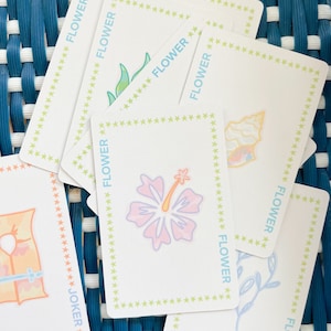 Pode incluir: Um baralho de cartas com uma borda de estrelas verde, amarela e branca. As cartas apresentam uma variedade de ilustra&ccedil;&otilde;es florais, incluindo um hibisco, uma concha e um ramo frondoso. As cartas tamb&eacute;m incluem uma carta de coringa com uma ilustra&ccedil;&atilde;o de salva-vidas.