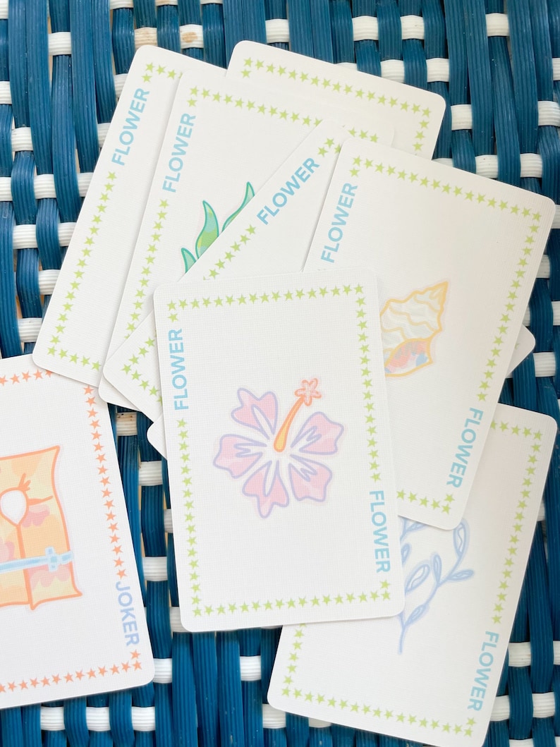 Pode incluir: Um baralho de cartas com uma borda de estrelas verde, amarela e branca. As cartas apresentam uma variedade de ilustra&ccedil;&otilde;es florais, incluindo um hibisco, uma concha e um ramo frondoso. As cartas tamb&eacute;m incluem uma carta de coringa com uma ilustra&ccedil;&atilde;o de salva-vidas.