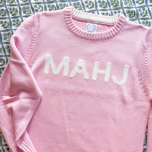 May include: A light pink sweater with the letters "MAHJ" in white across the front. The sweater has a ribbed crew neck and cuffs. The background is a blue and white floral pattern.