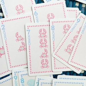 Pode incluir: Um conjunto de cartas de jogar rosa e azul com um design de caranguejo e estrelas. As cartas s&atilde;o numeradas de 1 a 9 e t&ecirc;m as palavras "Crack" e "Dot" nelas.