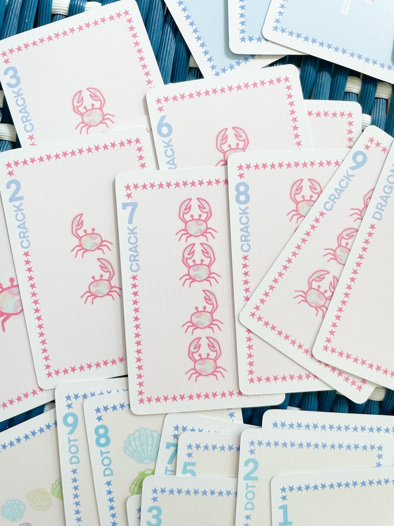 Pode incluir: Um conjunto de cartas de jogar rosa e azul com um design de caranguejo e estrelas. As cartas s&atilde;o numeradas de 1 a 9 e t&ecirc;m as palavras "Crack" e "Dot" nelas.