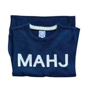 May include: Navy blue knit sweater with the white letters "MAHJ" across the front. The sweater has a ribbed crew neck with white trim and a small circular tag. The sweater is folded neatly.