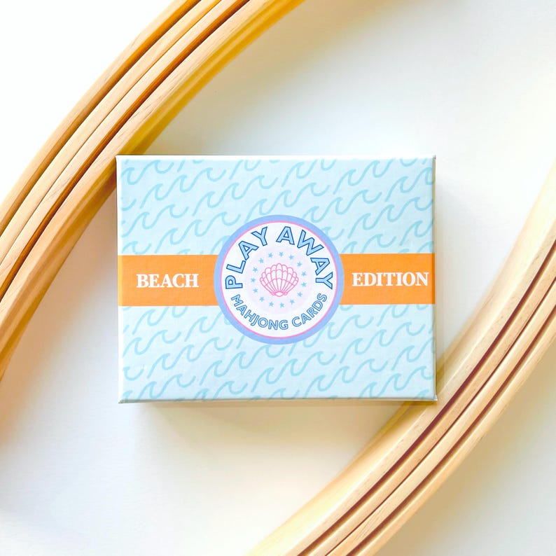 Pode incluir: Uma caixa azul com um c&iacute;rculo branco contendo o texto "Play Away Mahjong Cards" e uma concha rosa. A caixa tem uma faixa laranja claro com o texto "Beach Edition".