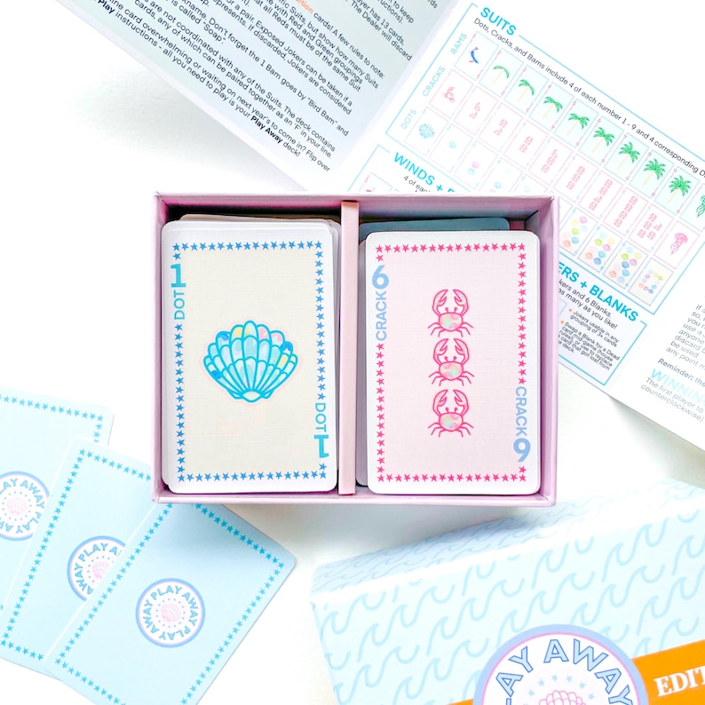 Pode incluir: Uma caixa rosa e azul de cartas de jogo com um design de concha e caranguejo. A caixa tem um fundo azul claro com um padr&atilde;o de ondas e o texto "Play Away" em um c&iacute;rculo.