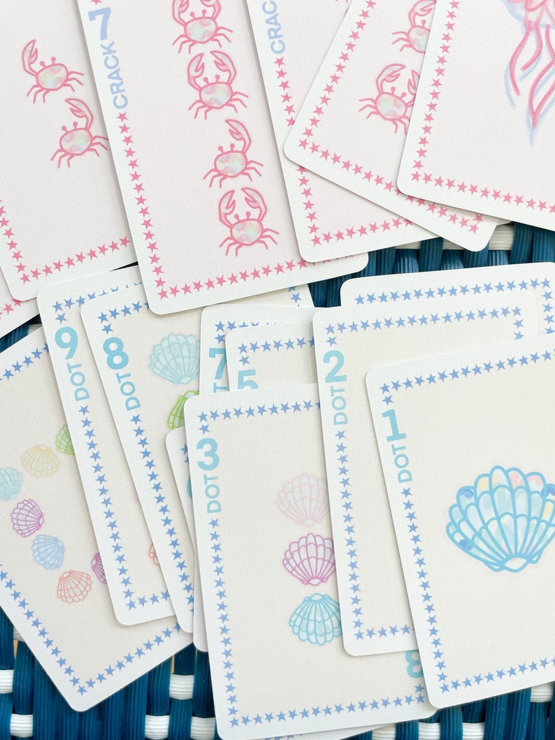 Pode incluir: Um conjunto de cartas de jogar coloridas com tema n&aacute;utico. As cartas apresentam imagens de conchas, caranguejos e as palavras "DOT" e "CRACK" com n&uacute;meros.