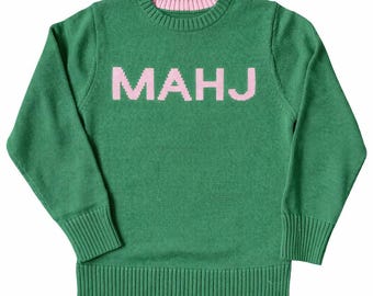 May include: Green knit sweater with the word "MAHJ" in pink block letters across the chest. The sweater has a ribbed hem and cuffs, and a pink trim around the neckline. The long-sleeved sweater is made of a soft knit material.