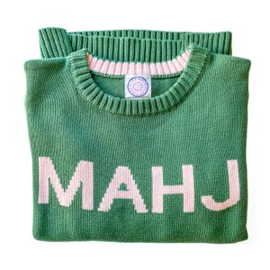 K&ouml;nnte beinhalten: Gr&uuml;ner Strickpullover mit dem Wort "MAHJ" in hellrosa Blockbuchstaben. Der Pullover hat einen gerippten Rundhalsausschnitt mit rosa Besatz und ein kleines Etikett. Der Pullover ist sauber auf wei&szlig;em Hintergrund gefaltet.