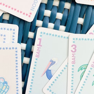 Pode incluir: Close-up de um jogo de cartas azul e rosa com uma ilustra&ccedil;&atilde;o de pelicano e o texto "BAM 1" no cart&atilde;o. O cart&atilde;o tem uma borda de estrelas azuis e rosas.