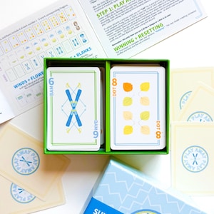 May include: A card game set with a green box containing two decks of cards. One deck features blue ski graphics, the other shows yellow and orange leaf illustrations. Additional cards and instructions are visible.