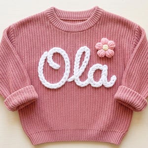 May include: A dusty rose long-sleeve jumper featuring a chunky knit texture, a ribbed crew neckline, and ribbed cuffs. The front displays the word Ola in white raised yarn lettering, accompanied by a small, soft pink flower applique.