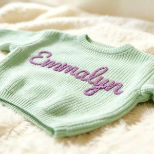 May include: A pale green knitted sweater featuring the name "Emmalyn" embroidered in purple script. The sweater has a ribbed texture and long sleeves. It is displayed on a white, fluffy surface, creating a soft aesthetic.