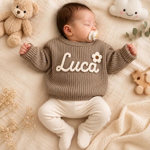 May include: A baby wearing a brown long-sleeved knit jumper with the name Luca embroidered in white script across the chest, accompanied by a small white flower appliqué. The baby is also wearing cream-coloured ribbed footed leggings.