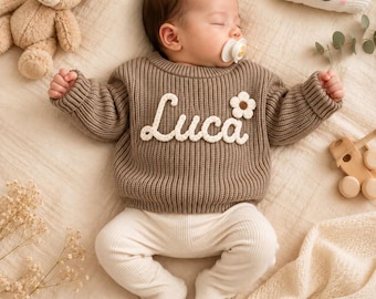 May include: A baby wearing a brown long-sleeved knit jumper with the name Luca embroidered in white script across the chest, accompanied by a small white flower appliqué. The baby is also wearing cream-coloured ribbed footed leggings.