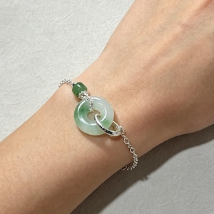 May include: A silver chain bracelet featuring a circular green jade donut pendant and a small green jade bead. The bracelet is secured with a silver link, showcasing a polished metallic finish and a smooth, translucent green stone design.