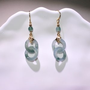 Könnte beinhalten: Ein Paar goldfarbene Ohrringe mit hellgrünem Jade-Design. Jeder Ohrring hat einen goldenen Haken, eine kleine grüne Perle, einen goldfarbenen blattförmigen Akzent und zwei ineinandergreifende Jade-Ringe. Die Ohrringe werden auf einer weißen Oberfläche präsentiert.