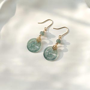 May include: A pair of gold-colored dangle earrings. Each earring features a round, pale green jade pendant with a carved design, a small green bead, and a gold-colored charm. The earrings hang from gold-colored ear wires.