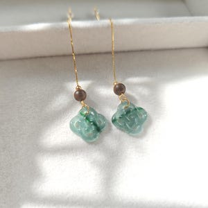 May include: A pair of green jade earrings with a gold chain and a brown wooden bead. The jade is carved into a knot design.