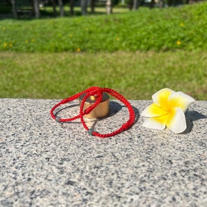 May include: A red string bracelet with two grey beads and a gold metal charm. The bracelet is lying on a grey stone surface with a white flower.