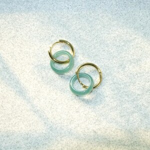 May include: Two pairs of gold hoop earrings with a light green, circular jade stone. The earrings are displayed on a white, textured surface. The earrings are a simple, elegant design.