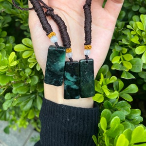 Könnte beinhalten: Drei Jade-Anhänger an braunen Schnüren mit goldenen Perlen. Die Anhänger sind rechteckig und haben eine dunkelgrüne Farbe mit weißen Streifen.