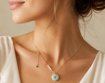 May include: A gold chain necklace featuring a pale green circular jade pendant, a small cluster of four white pearls, and a dangling accent chain finished with a single small green gemstone. The necklace is worn around the neck.