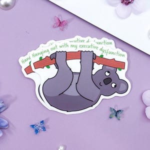 May include: A sticker featuring a cartoon koala hanging from a branch. The koala is gray with a white belly and black eyes. Text on the sticker reads "Hang Hanging out with my executive dysfunction."