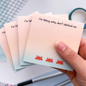 May include: A stack of square sticky notes with a gradient pink and blue background. The notes have the text "I'm taking notes, don't sidetrack me" and feature red crab illustrations. A hand holds the notes.