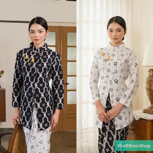 Op de afbeelding: Twee vrouwen in traditionele Indonesische batik outfits. De ene outfit is zwart-wit met een geometrisch patroon, de andere wit met een bloemenontwerp. Beide outfits hebben een bijpassende rok en een decoratieve broche.