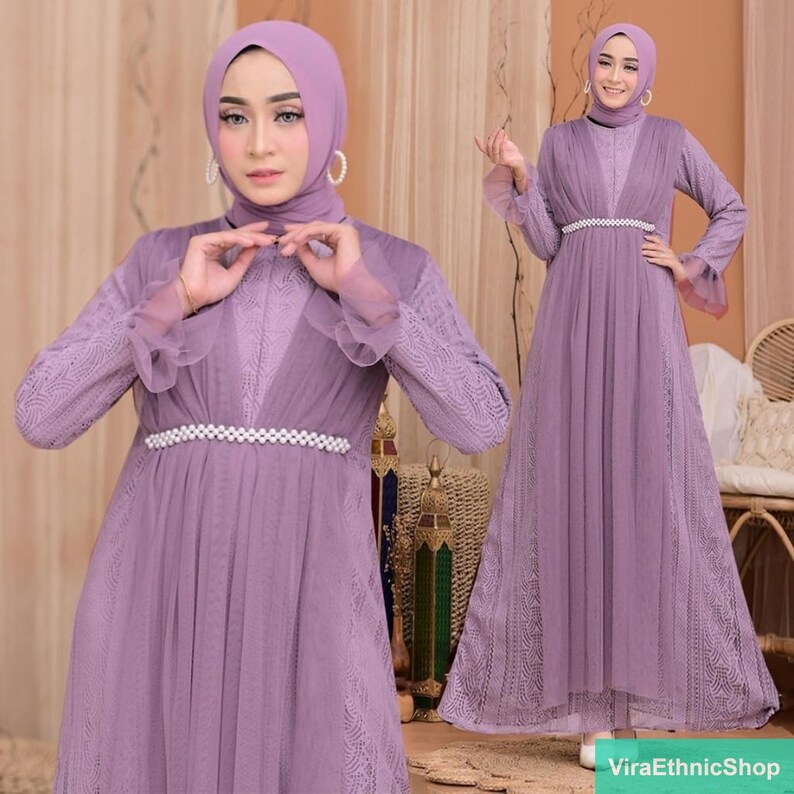 May include: A long, lilac-colored dress with a lace overlay and a pearl-embellished belt. The dress has long sleeves with ruffles at the cuffs. The model is wearing a matching lilac headscarf.